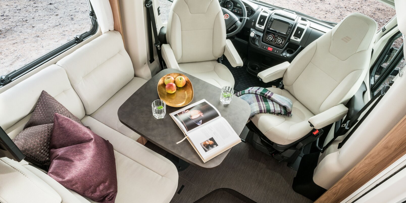 Spazio abitativo dell’autocaravan HYMER Exsis-t con sedili cabina guida, tavolo apparecchiato con piatto con frutta, libro e bicchieri