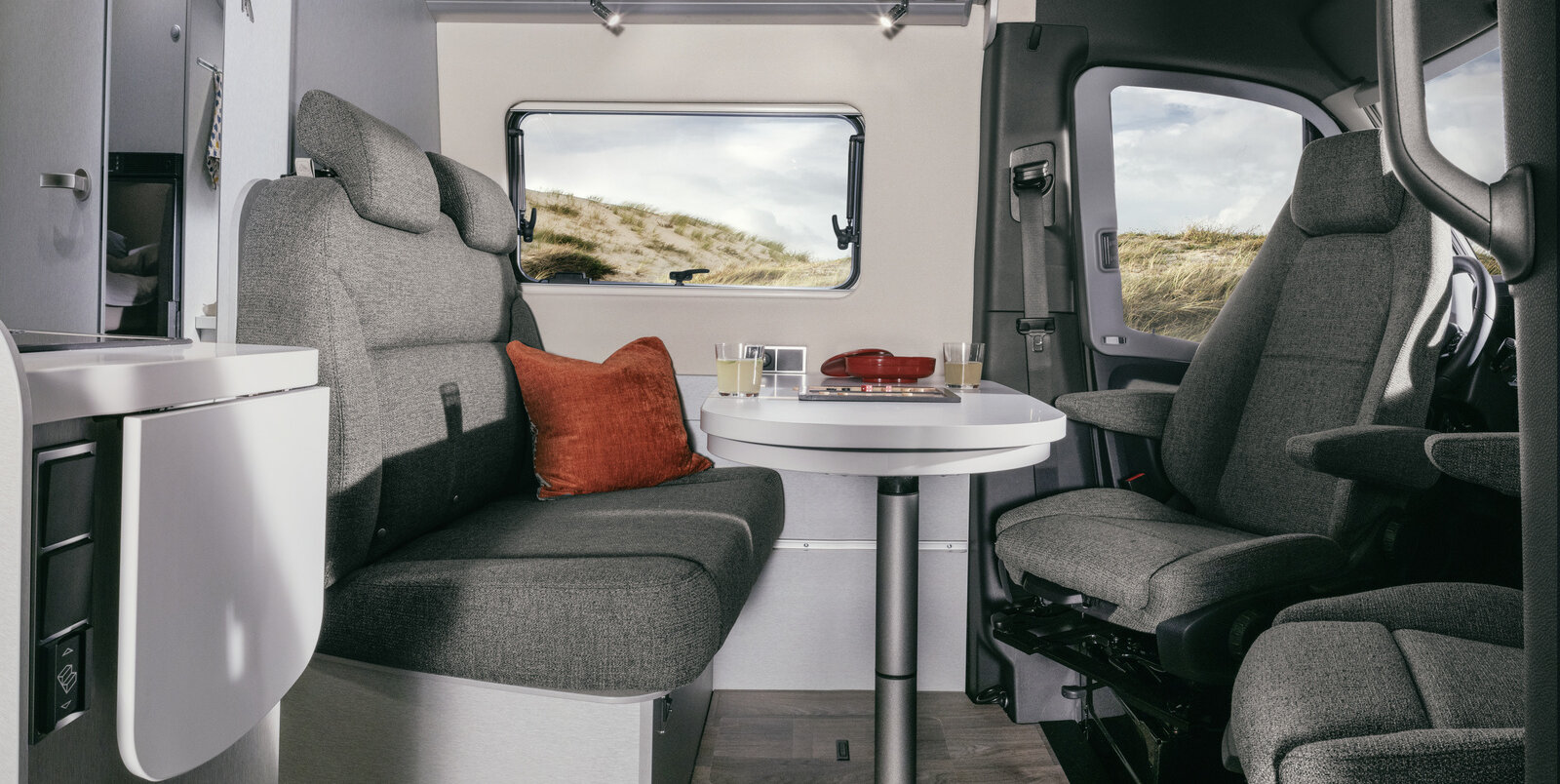 Seating group with driver's seats, occupied table, red decorative cushions on bench and overhead locker in the HYMER Camper Van Free S 600