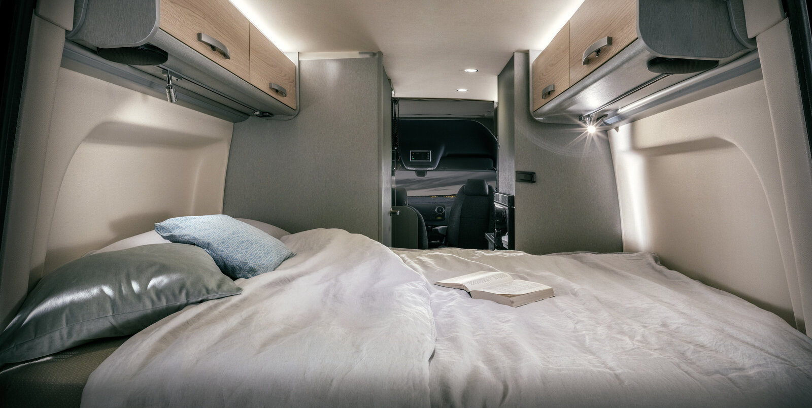 Cama cubierta con almohadas, libro abierto, luces de lectura, armarios superiores en la parte trasera del HYMER Free S 600