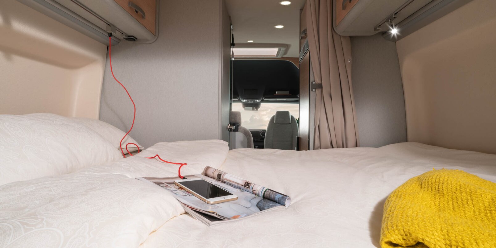 Opgemaakt bed met gele vlonder, tijdschrift, mobiele telefoon met ingeplugde laadkabel en leeslampjes in de HYMER Free S 600