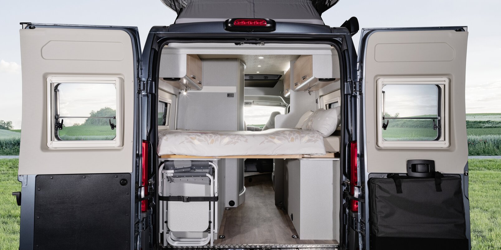 Porte posteriori aperte, letto con lenzuola, sedie da campeggio sistemate, visuale interna fino alla cabina guida del Free HYMER in un prato