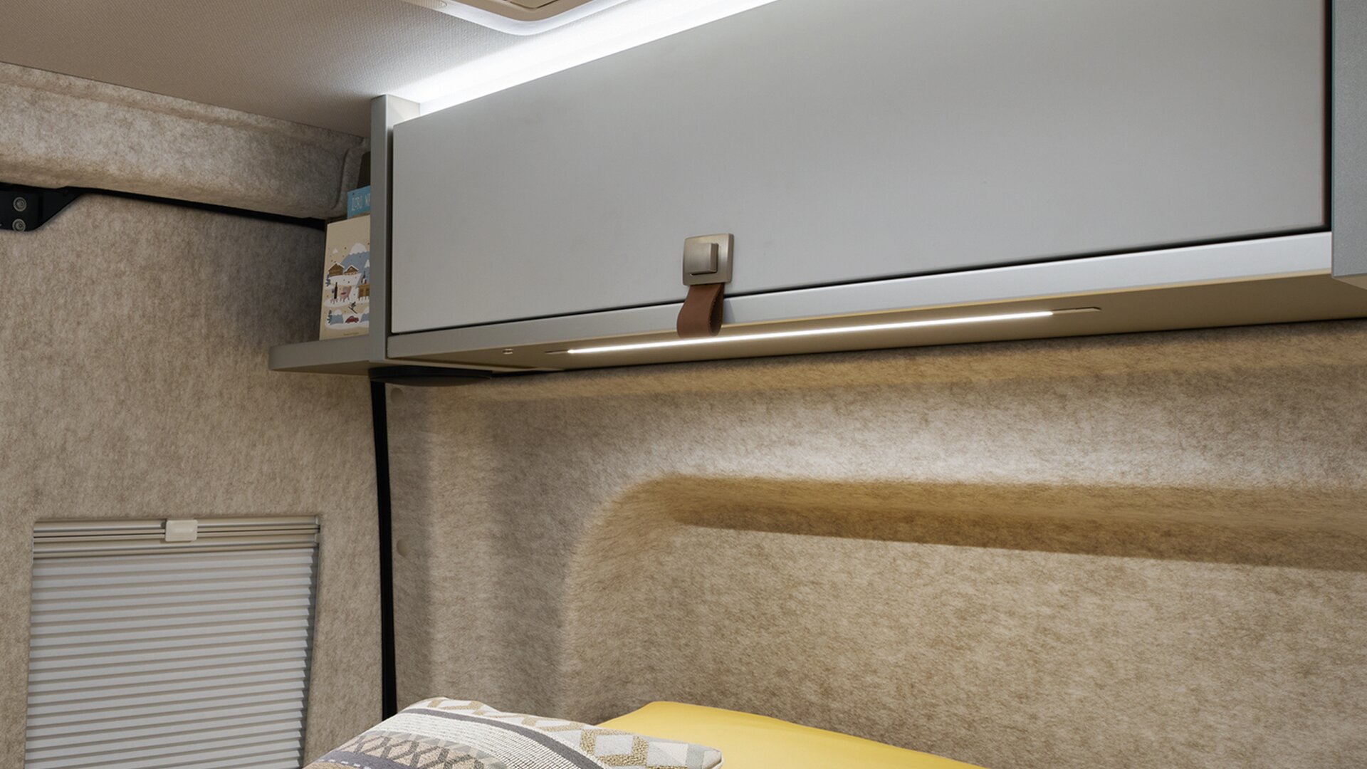 Le pareti del HYMER Grand Canyon S nel mondo dello stile Pearl Grey sono rivestite in feltro. La luce sopra gli armadietti sul tetto è accesa.