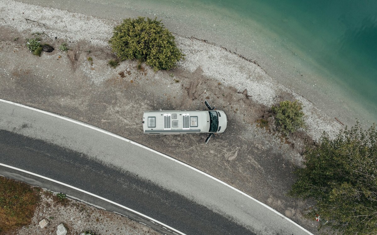Grey camper van from above, standing on gravel next to a lake at the shore, next to the gravel there is a small road and on the gravel bed there are three green trees, the doors of the camper van are open