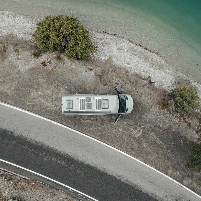 Furgone grigio dall'alto, parcheggiato su ghiaia vicino a un lago sulla riva, accanto alla ghiaia c'è una piccola strada e sul letto di ghiaia ci sono tre alberi verdi, le porte del furgone sono aperte