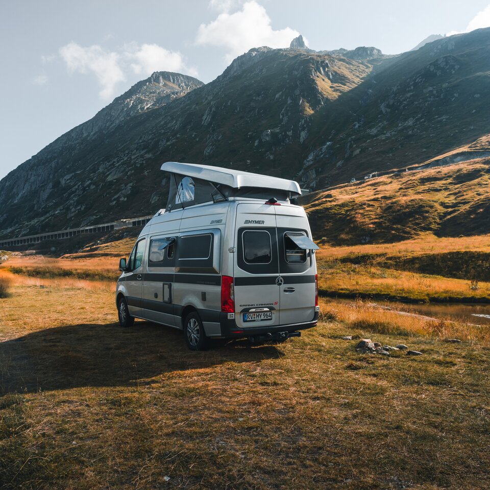 Der HYMER Grand Canyon S ist von schräg hinten zu sehen. Er steht leicht eingedreht vor einem Bergpanorama auf einer Wiese.