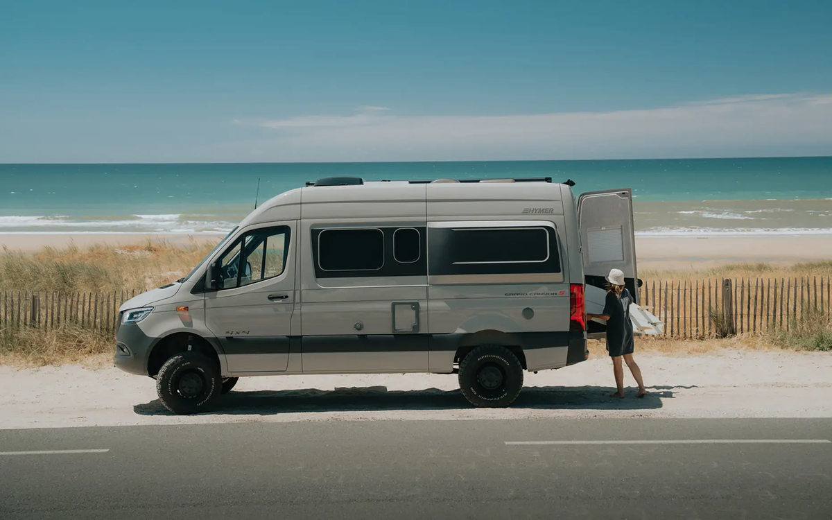 Vista laterale di un camper van grigio pietra con accenti di design neri e pneumatici all-terrain neri, parcheggiato sul ciglio della strada direttamente sulla spiaggia con il mare turchese sullo sfondo sotto un cielo azzurro brillante. Una persona è in piedi alla porta posteriore aperta con una tavola da surf in mano.
