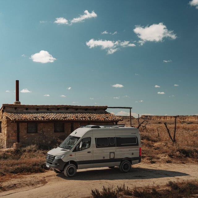 Campervan grigio chiaro con accenti di design nero è parcheggiato lateralmente davanti a un edificio in pietra a un piano in un paesaggio arido, simile al deserto sotto un cielo azzurro con pochissime piccole nuvole