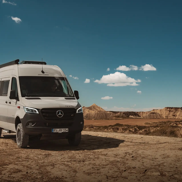 Il campervan grigio chiaro di HYMER è leggermente inclinato con la parte anteriore rivolta in avanti in un paesaggio arido, simile al deserto sotto un cielo azzurro con poche nuvole bianche