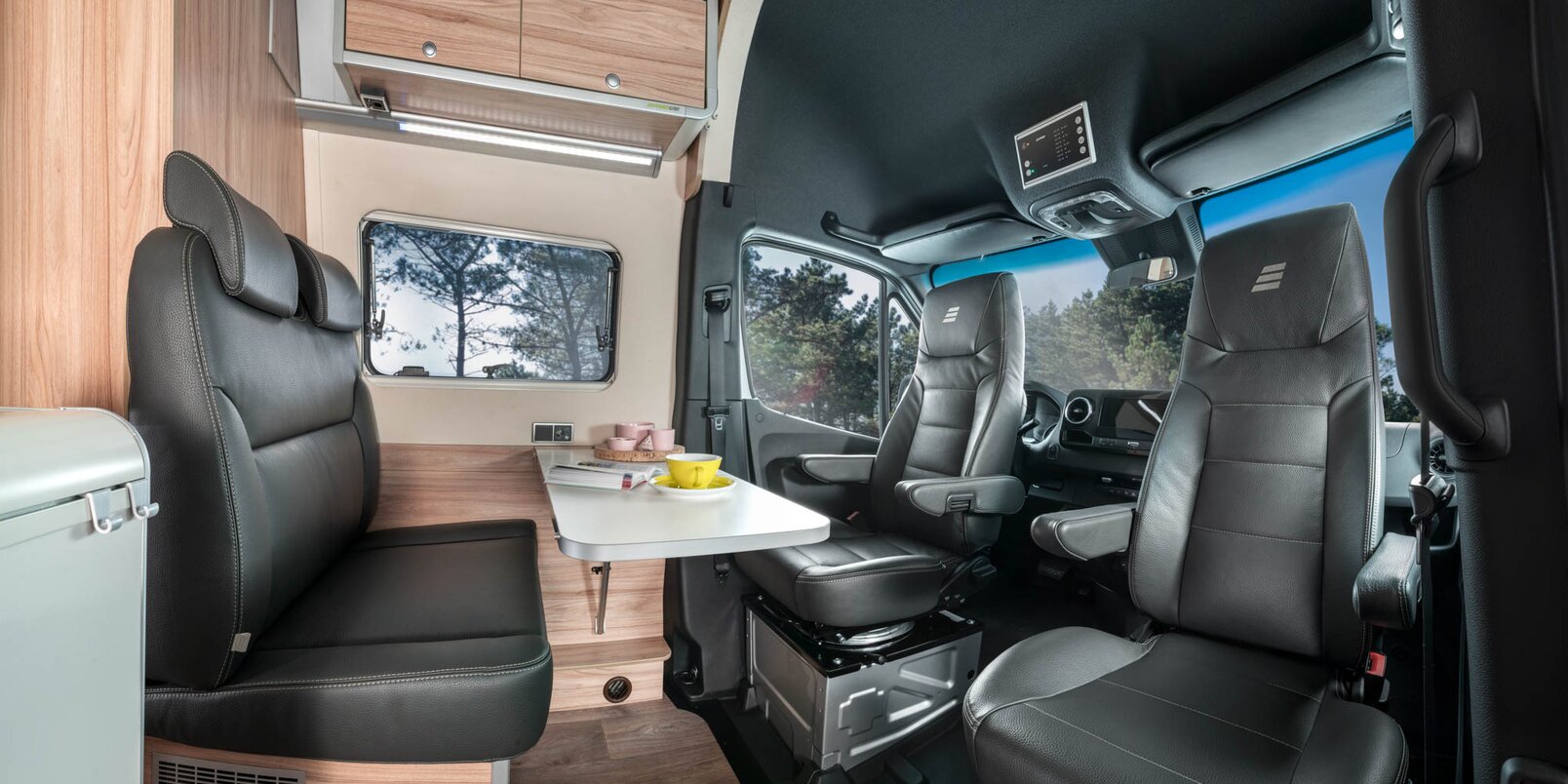 Seating group with driver and front passenger seats, table with yellow mug, magazine and overhead storage cupboards in the HYMER Grand Canyon S