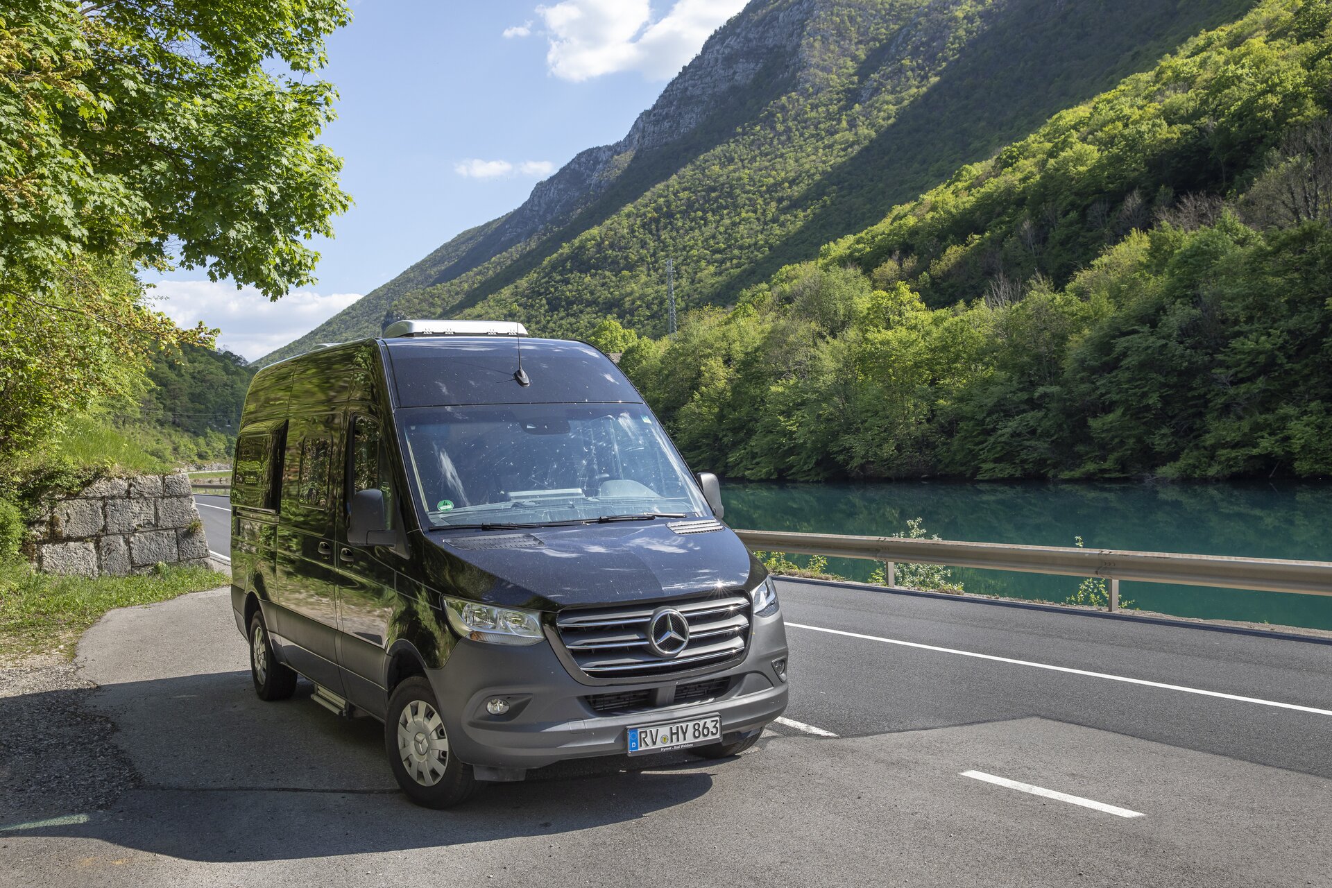 Der schwarze Mercedes Campingbus HYMER Grand Canyon S steht auf einer Straße neben einem Fluss in bergiger Landschaft, auf den Bergen wachsen Bäume.