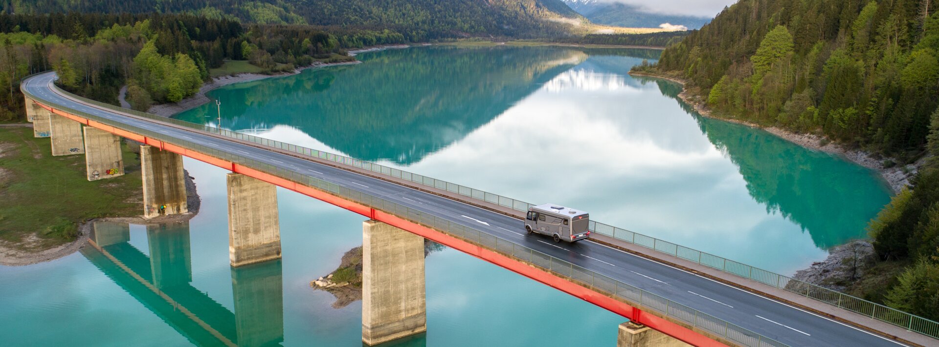 HYMER B-Class ModernComfort I on a bridge with concrete pillars, turquoise lake, surrounded by forest