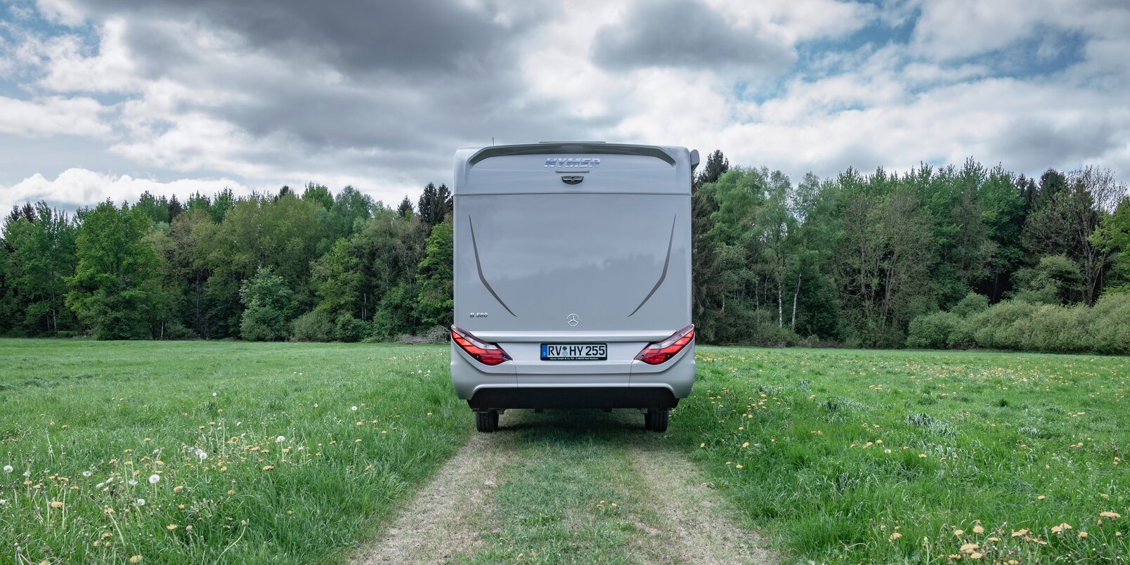 Rear of the HYMER B-MC on a meadow path with dandelions and forest