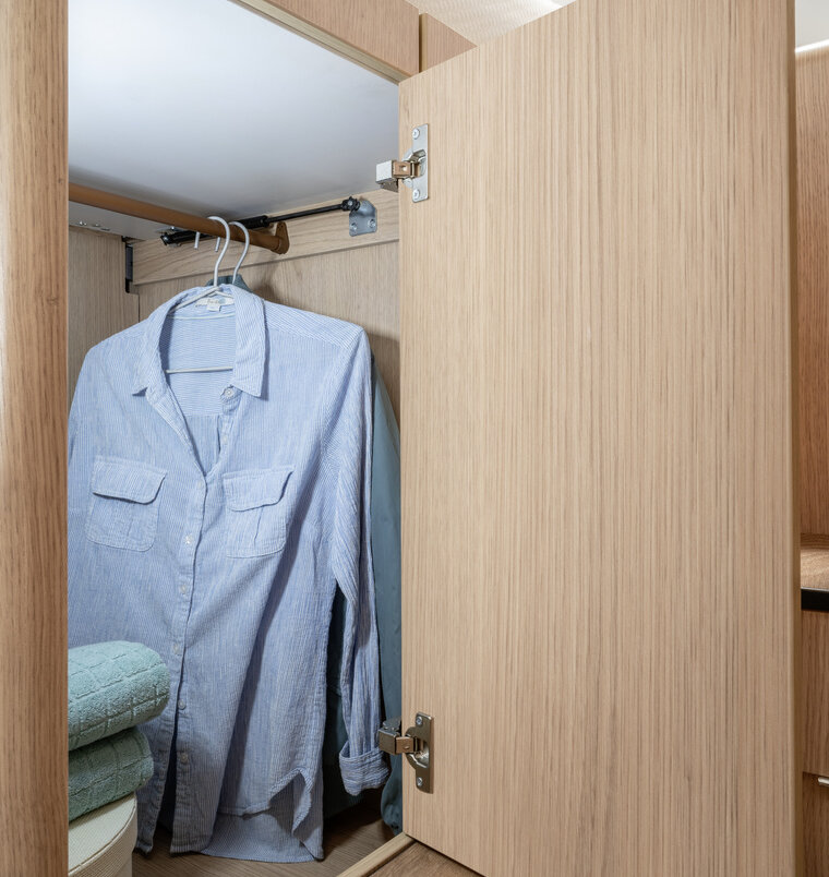 Geöffneter, beleuchteter Kleiderschrank mit Hemden auf der Stange und Handtüchern unter dem Bett im Heck des HYMER B-MC I