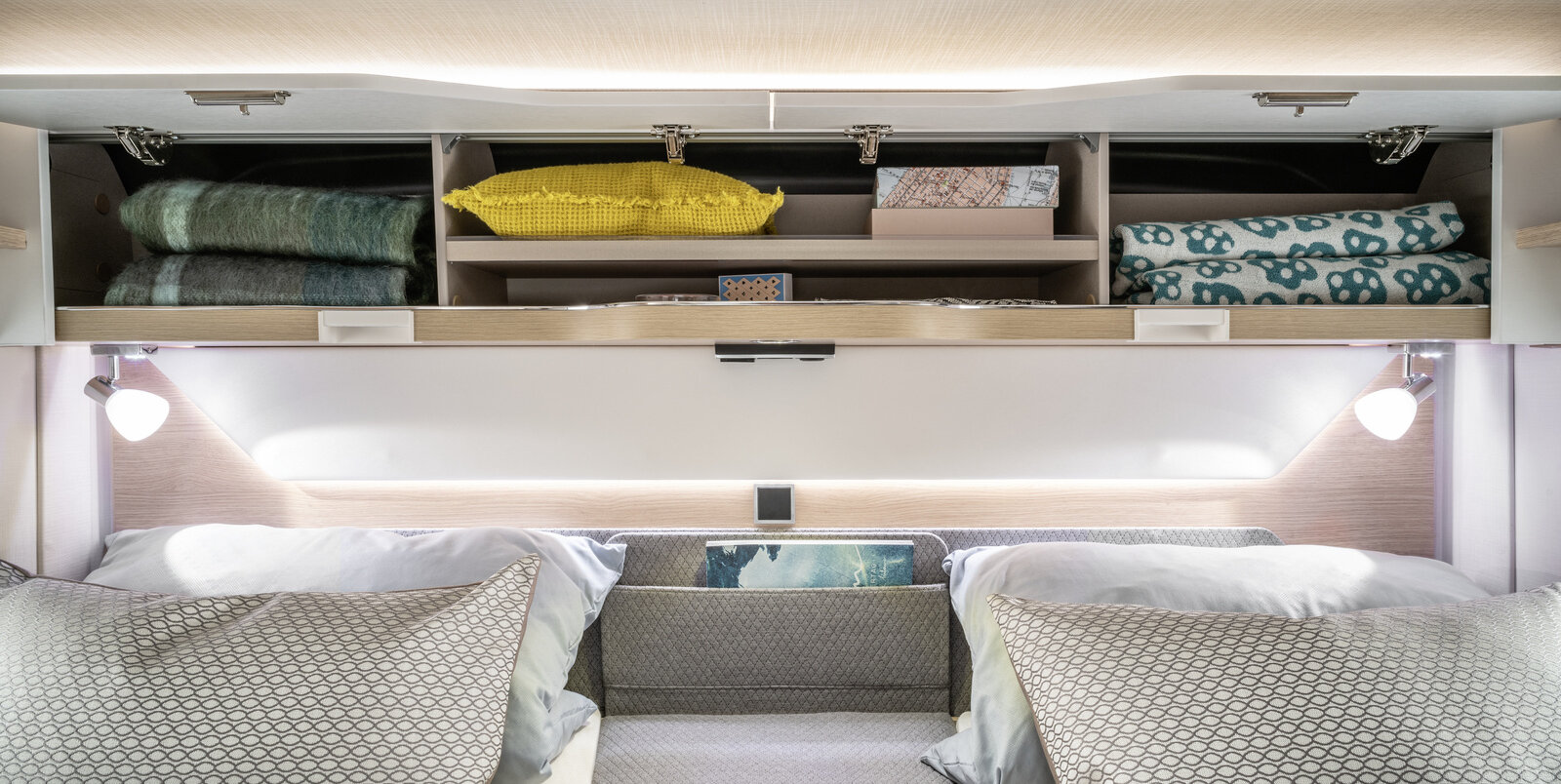 Open bovenkasten gevuld met dekens over het bed achterin het voertuig, bedverlichting en kussens in de Hymer B MC