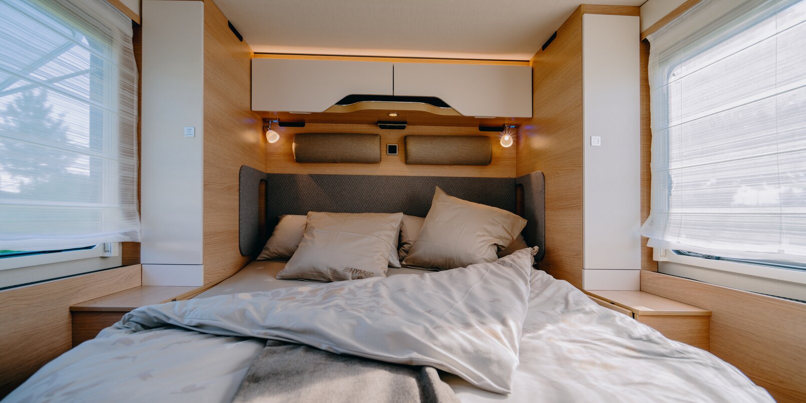 Schlafbereich: Mit Bettwäsche bezogenes Queensbett, Dachstauschränke und seitlich je ein langer Schrank im HYMER B-MC T 690