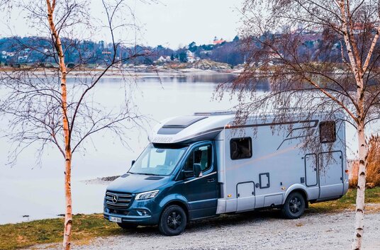 HYMER B-Class ModernComfort T on a lake between two birch trees and a small settlement on the other bank
