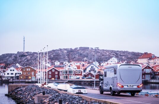 Rear view of the HYMER B-MC T at a small harbor with the typical Scandinavian wooden houses and rock face behind them