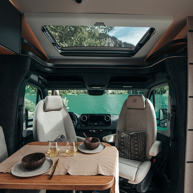 Der Tisch des HYMER ML-T in der Stilwelt Native Bamboo ist gedeckt mit Geschirr und Weißwein. Dahinter liegt das Fahrerhaus mit Blick in die Natur.
