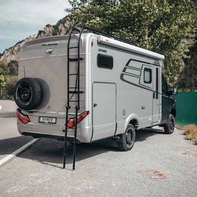Der HYMER ML-T ist von schräg hinten zu sehen. Er steht leicht eingedreht vor einem Gebirgssee und die Dachleiter ist ausgefahren.