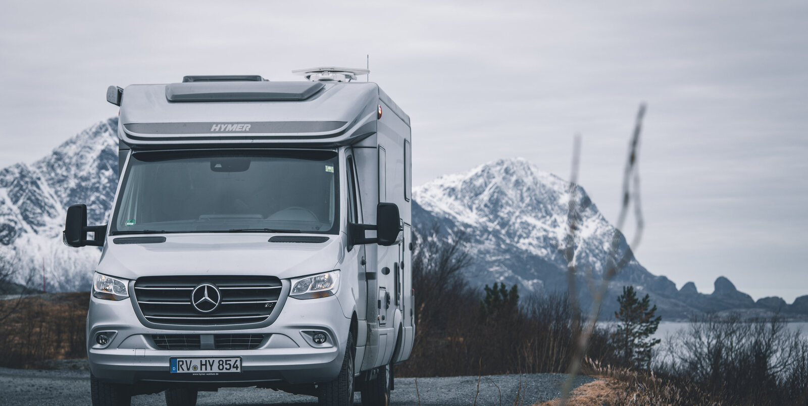 HYMER ML-T motorhome in front of snow-covered mountains and a lake on a gravel road with grasses / bushes on the hard shoulder