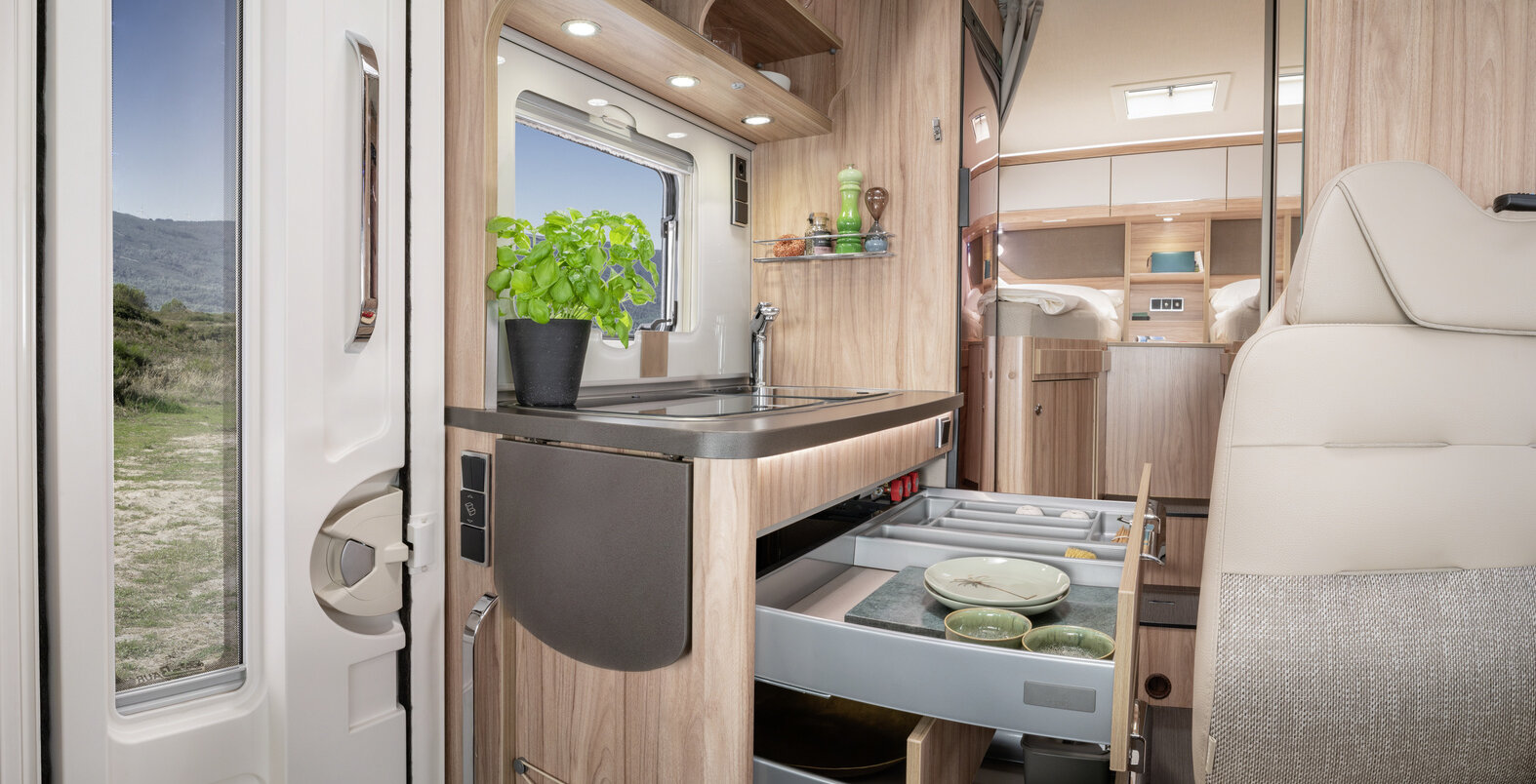 Entrance door, kitchen  with open drawers filled with dishes, cutting board and herb pot on shelf in the HYMER ML-T