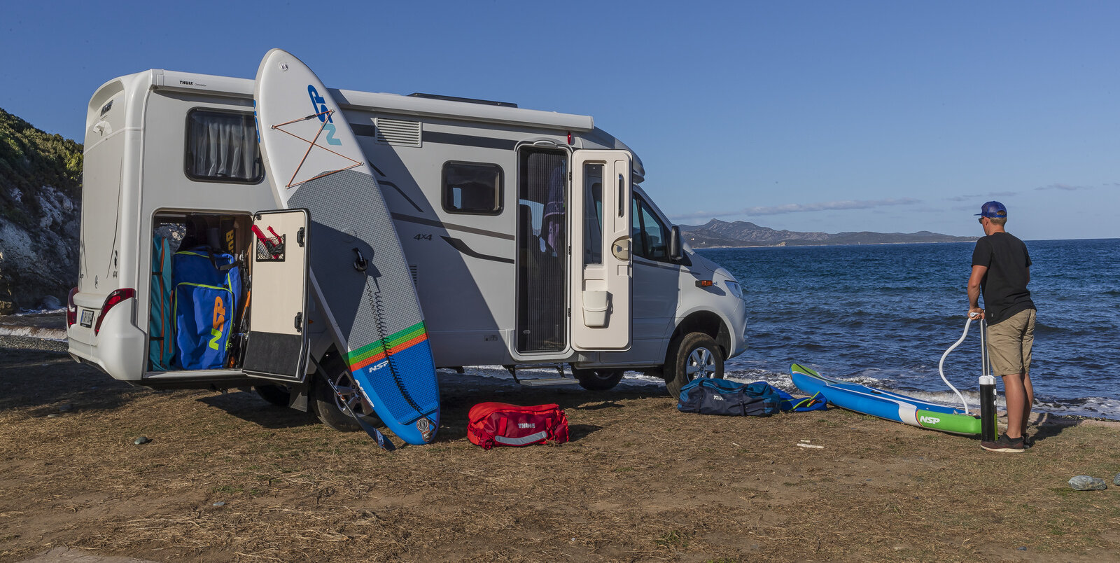 Two people on the beach with a surfboard leaning against the HYMER ML-T and a rear garage filled with sports equipment