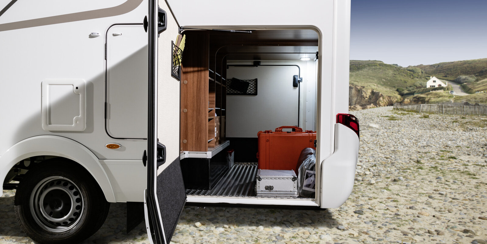 Opened rear garage of the HYMER ML-T loaded with red and silver suitcases in a landscape with sand, grass and house