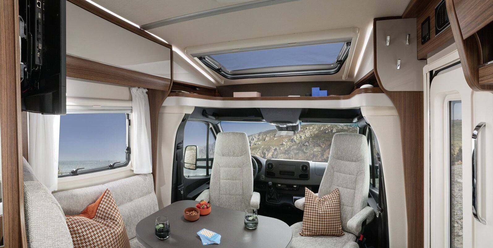 Living room in the HYMER ML-T: seating area with cushions, laid table, entrance door, panoramic roof vent, driver's seats, storage board