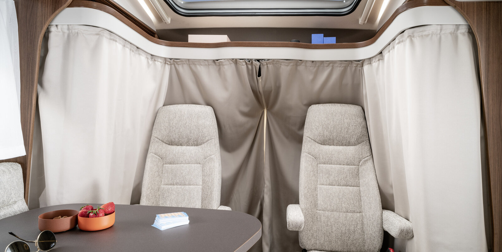 Driver's cab with seats, curtains all around as a privacy screen, table with sunglasses, two bowls and playing cards in the HYMER ML-T