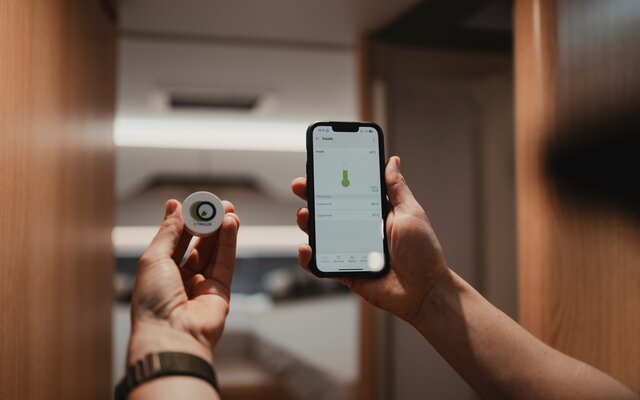 Hands of a person holding a temperature sensor in the left hand and a smartphone using the Connect app in the right hand measuring the current temperature, with the sleeping area of a motorhome blurred in the background