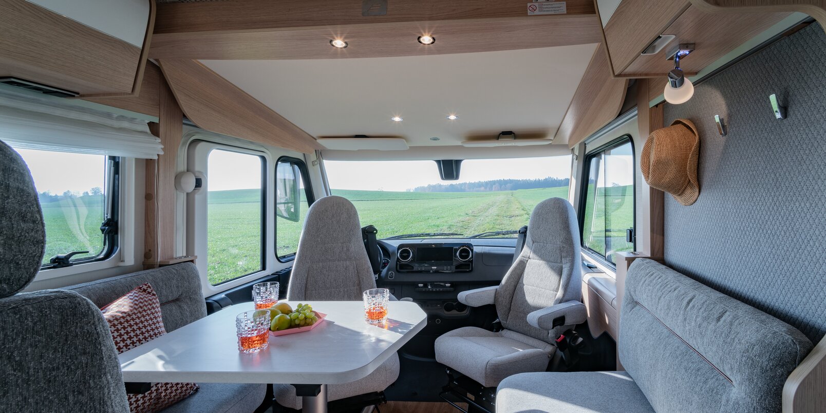 Woonkamer in de HYMER B-MC I 550 WL, bestuurdersstoelen, gedekte tafel, geruite sierkussens, uitzicht op de natuur door het raam