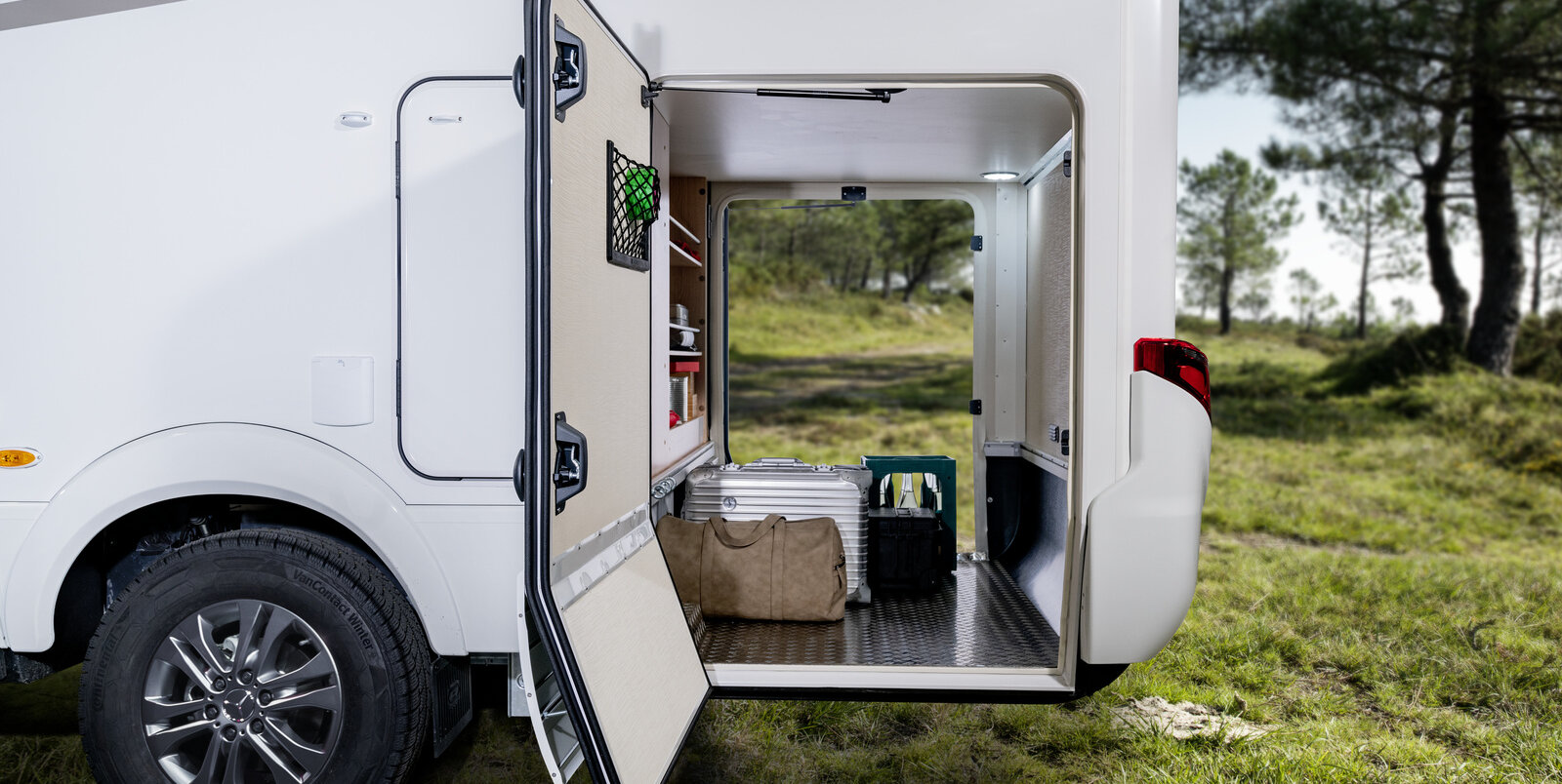 Opened doors of the rear garage of the HYMER B-MC loaded with suitcase, water box and luggage on a meadow lined with forest