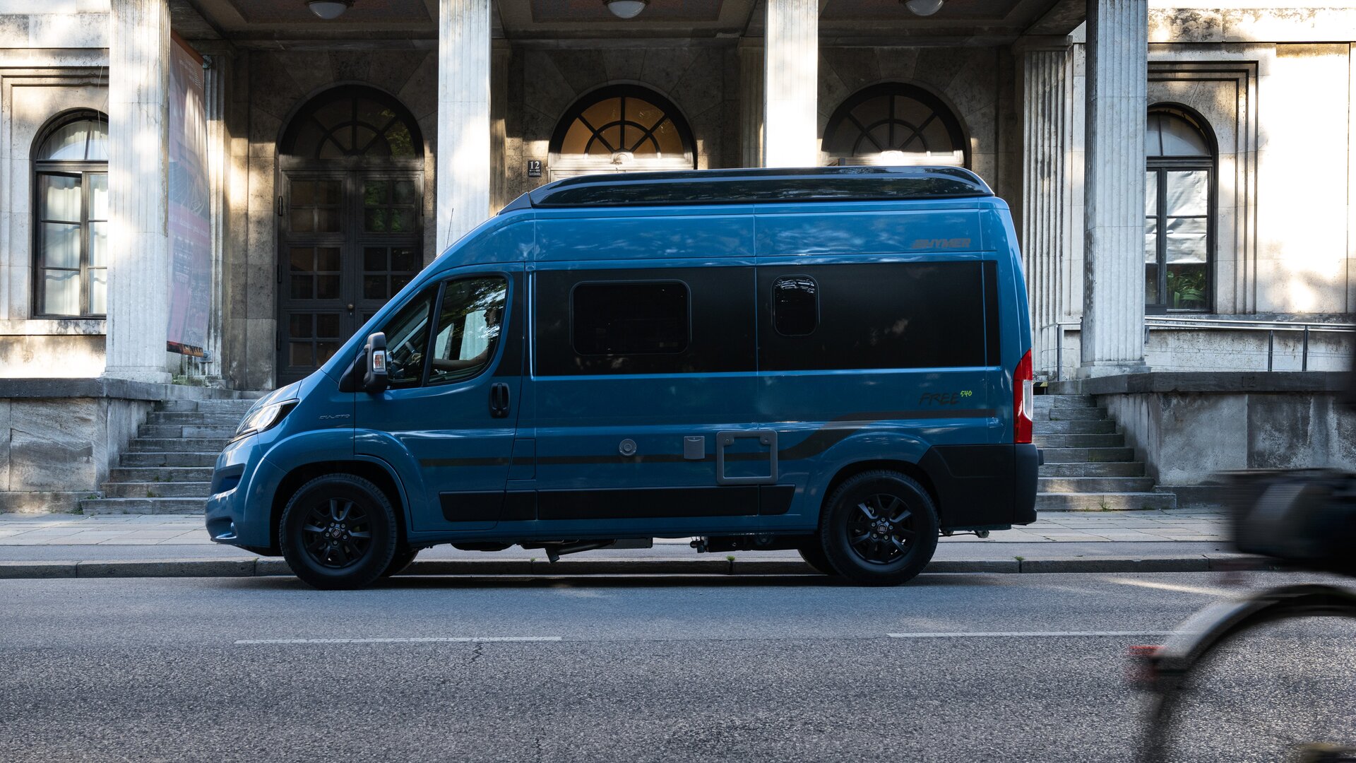 HYMER Free 540 Blue Evolution, painted blue metallic, drives past an old building