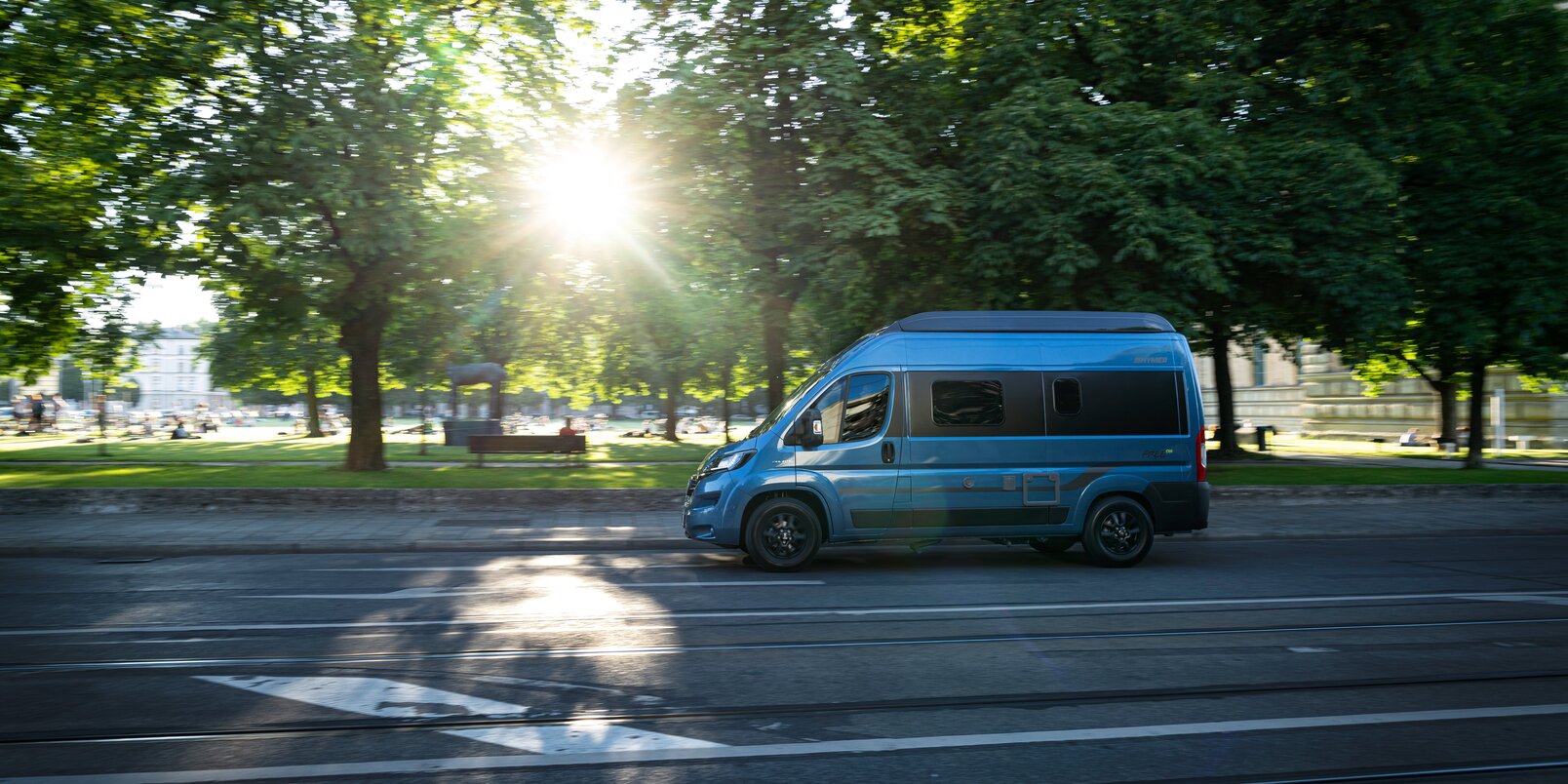 HYMER Free 540 Blue Evolution driving on a road next to a park with meadow, trees, benches and people