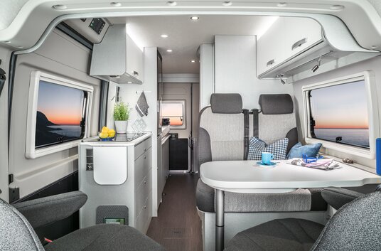 Interior in the HYMER Free 600 Campus: seating area with table, entrance door, kitchen block, side window, overhead storage cupboards