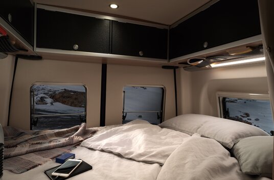 Cama con funda, almohadas, manta, teléfono móvil, portátil y armarios  superiores en la parte trasera del HYMER Grand Canyon S CrossOver