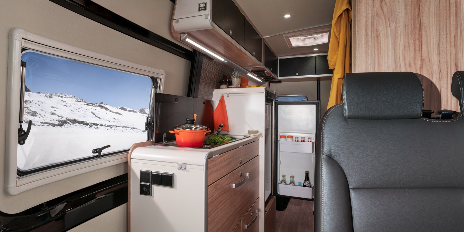Bench, kitchen block with open refrigerator, overhead storage cupboards, window with mountain view in the HYMER Grand Canyon S CrossOver