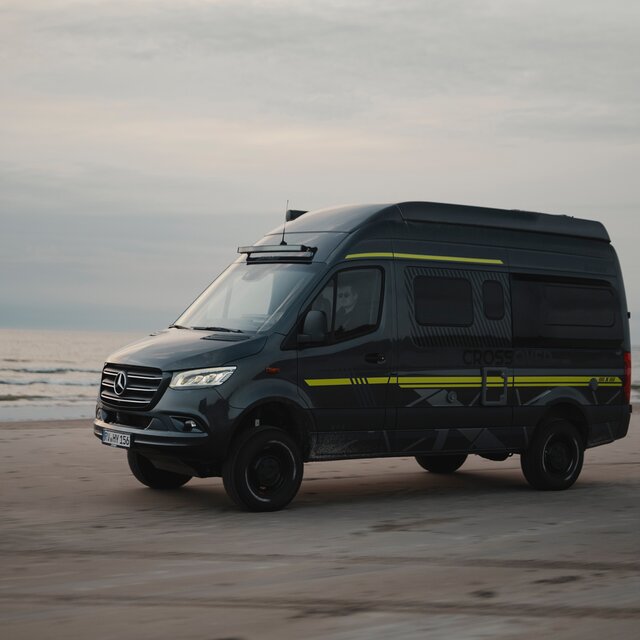 Schwarzer Camper Van mit Neongrüner Sonderbeklebung steht seitlich am Strand, im Hintergrund sieht man das Meer