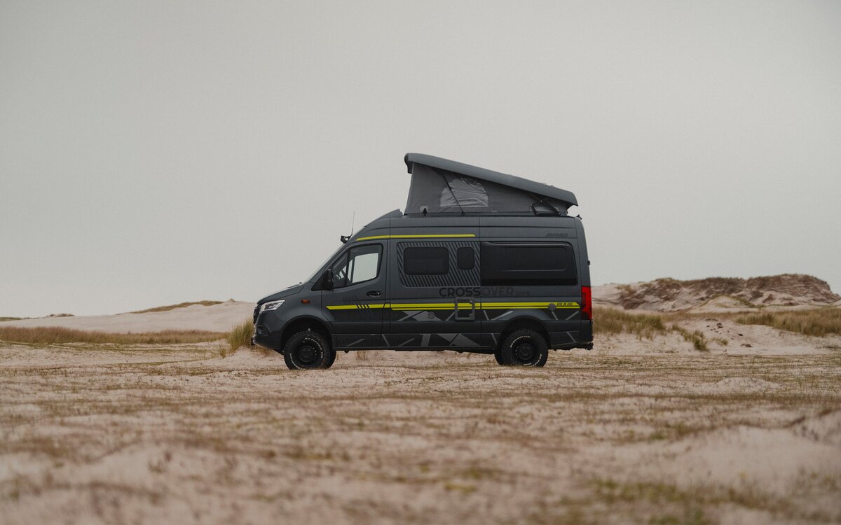 Schwarzer Camper Van mit Neongrüner Sonderbeklebung steht seitlich im Sand, im Hintergrund sieht man Dünen, das Aufstell Schlafdach ist offen