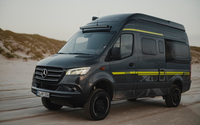 Schwarzer Camper Van mit Neongrüner Sonderbeklebung fährt über Sand, im Hintergrund sieht man Dünen