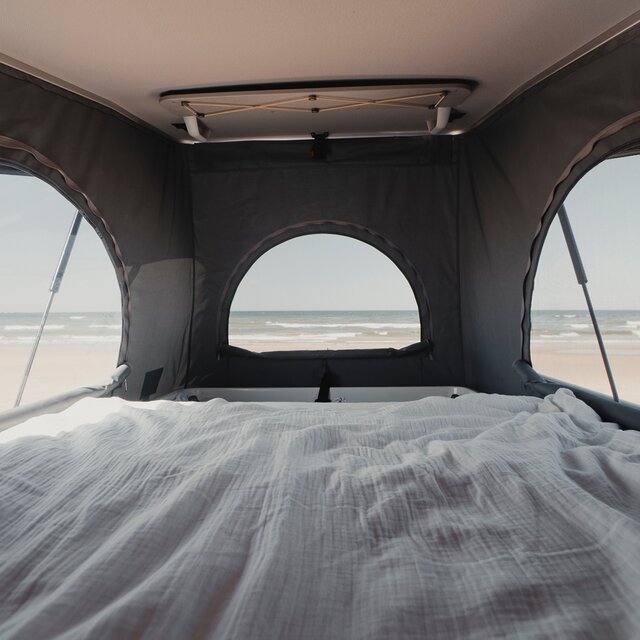 Schlafdach eines Camper Vans von innen, mit gemachtem Bett und Blick durch die geöffneten Front- und Seitenfenster auf den Strand und das Meer