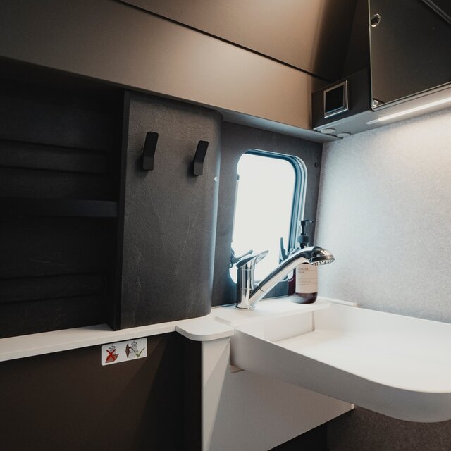 Waschbecken in einem Badezimmer in einem Camper Van mit silbernem Wasserhahn, links daneben sieht man ein Stauregal in dunkelgrauer Marmor Optik und zwei schwarze Handtuchhaken