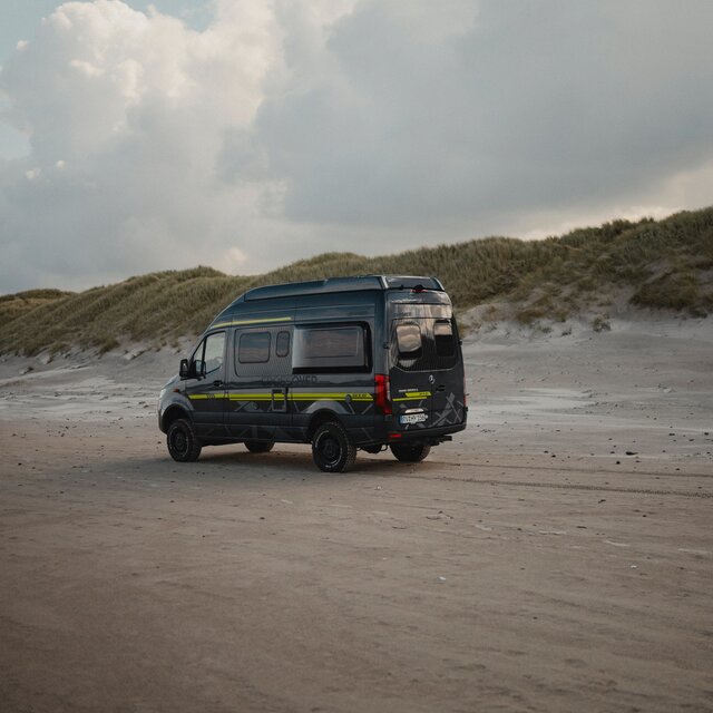 Schwarzer Camper Van mit Neongrüner Sonderbeklebung fährt über Sand, im Hintergrund sieht man Dünen