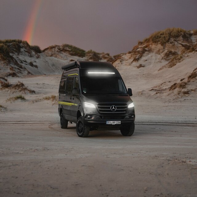 Schwarzer Camper Van mit Neongrüner Sonderbeklebung steht im Sand, im Hintergrund sieht man Dünen, die Scheinwerfer und die Lightbar leuchten in der Dämmerung