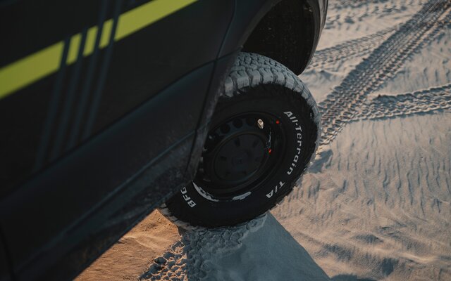 Schwarzer Reifen mit weißer All-Terrain T/A Beschriftung an einem dunklen Camper Van der auf Sand steht