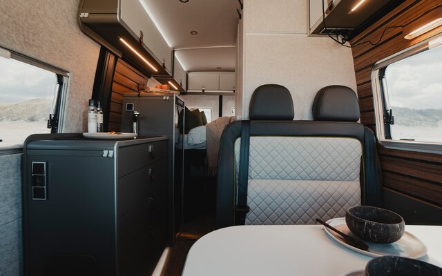 Wohnraum in einem modernen Camper Van, gedeckter Tisch und Sitzgruppe mit dunklem Leder, Küchenzeile mit Kochutensilien, Kühlschrank und Dachstauschränken, im Hintergrund sieht man ein gemachtes Bett