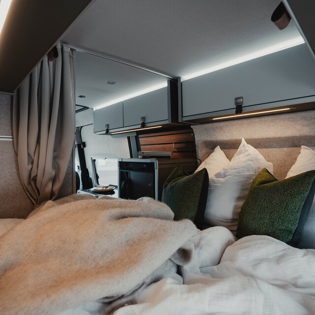 Schlafbereich in einem Camper Van, bezogenes Bett, zwei grüne Dekokissen und helle Dachstauschränke, im Hintergrund sieht man den Kühlschrank der Küchenzeile
