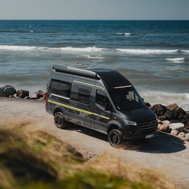 Schwarzer Camper Van mit grüner Beklebung steht seitlich neben großen Steinen und direkt dahinter sieht man das Meer