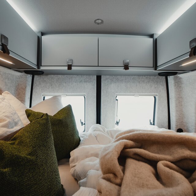 Schlafraum in einem Camper Van mit gemachtem Bett, mit grünen Dekokissen und hellen Dachstauschränken