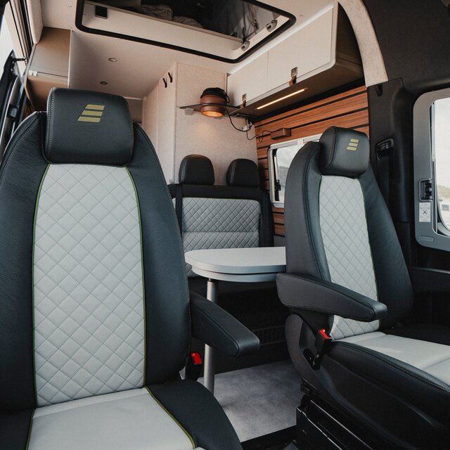 Zwei Fahrersitze in einem Camper Van mit schwarz grauem Lederbezug, im Hintergrund sieht man die gleich farbige Sitzgruppe mit dem davor stehenden Tisch und darüber die hellen Dachstauchschränke sowie die braune Pendelleuchte